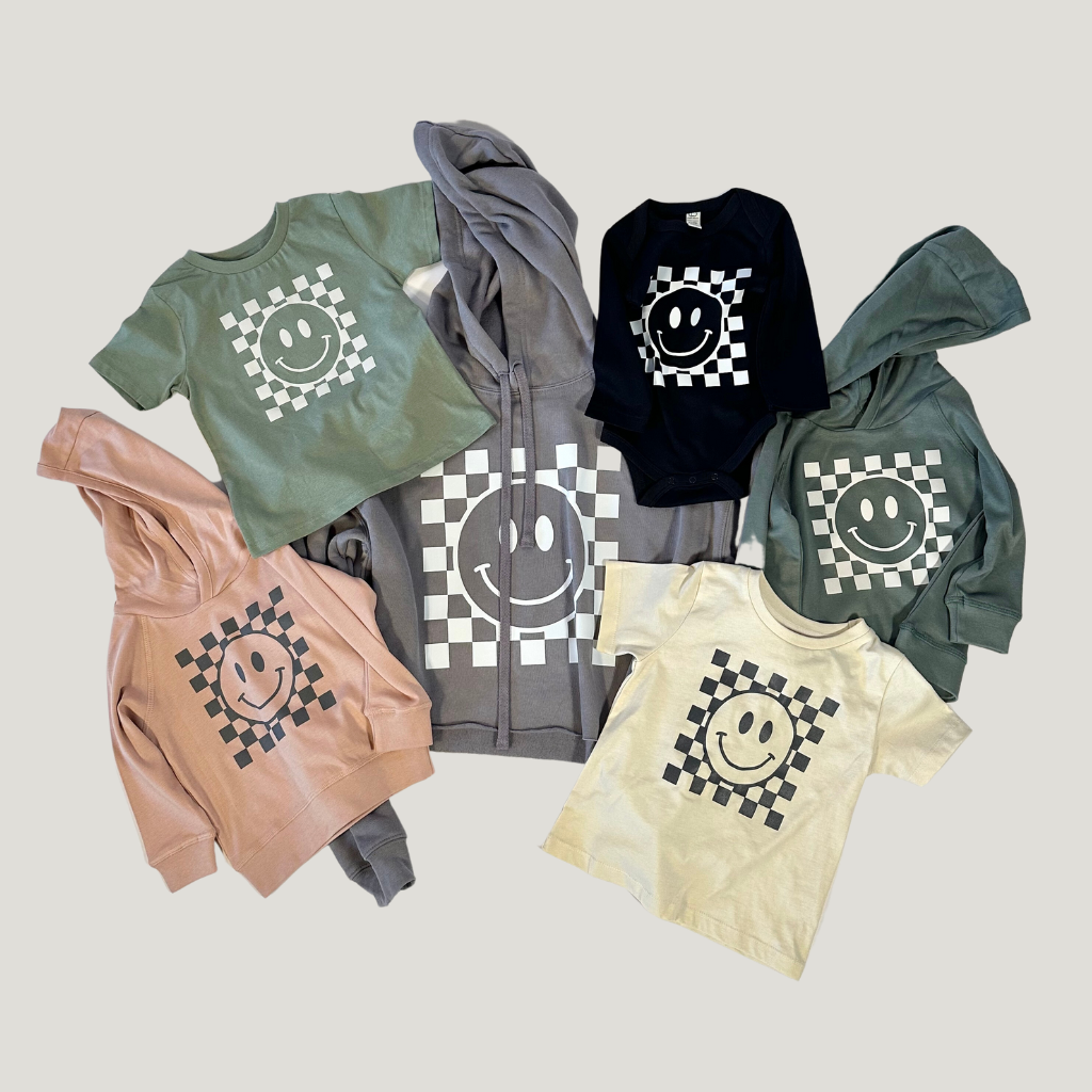 Assorted children's apparel laid out featuring a checkered smiley face design, including olive, black, and grey hoodies, along with cream and tan t-shirts