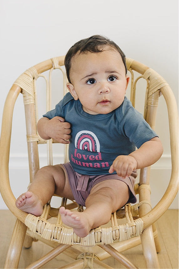 Curious baby in a slate blue 'loved human' onesie seated in a woven rattan chair, with a contemplative expression