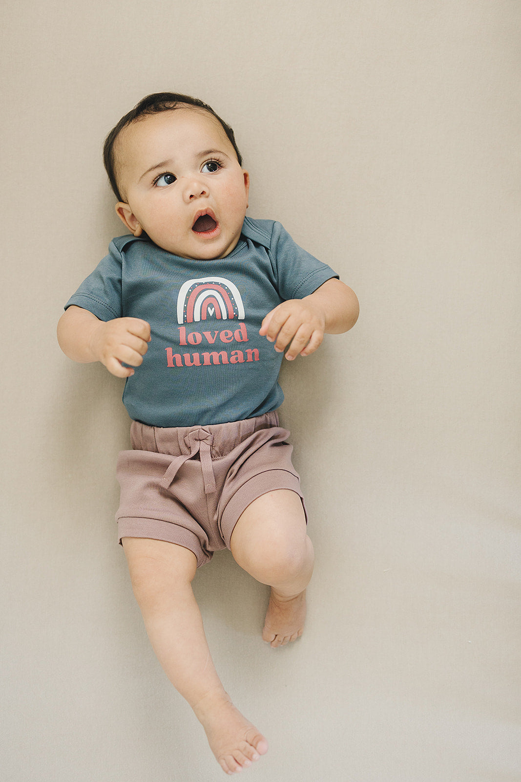 Adorable baby in a slate grey 'loved human' rainbow onesie with dusty rose shorts, lying on a beige background, looking surprised and delighted