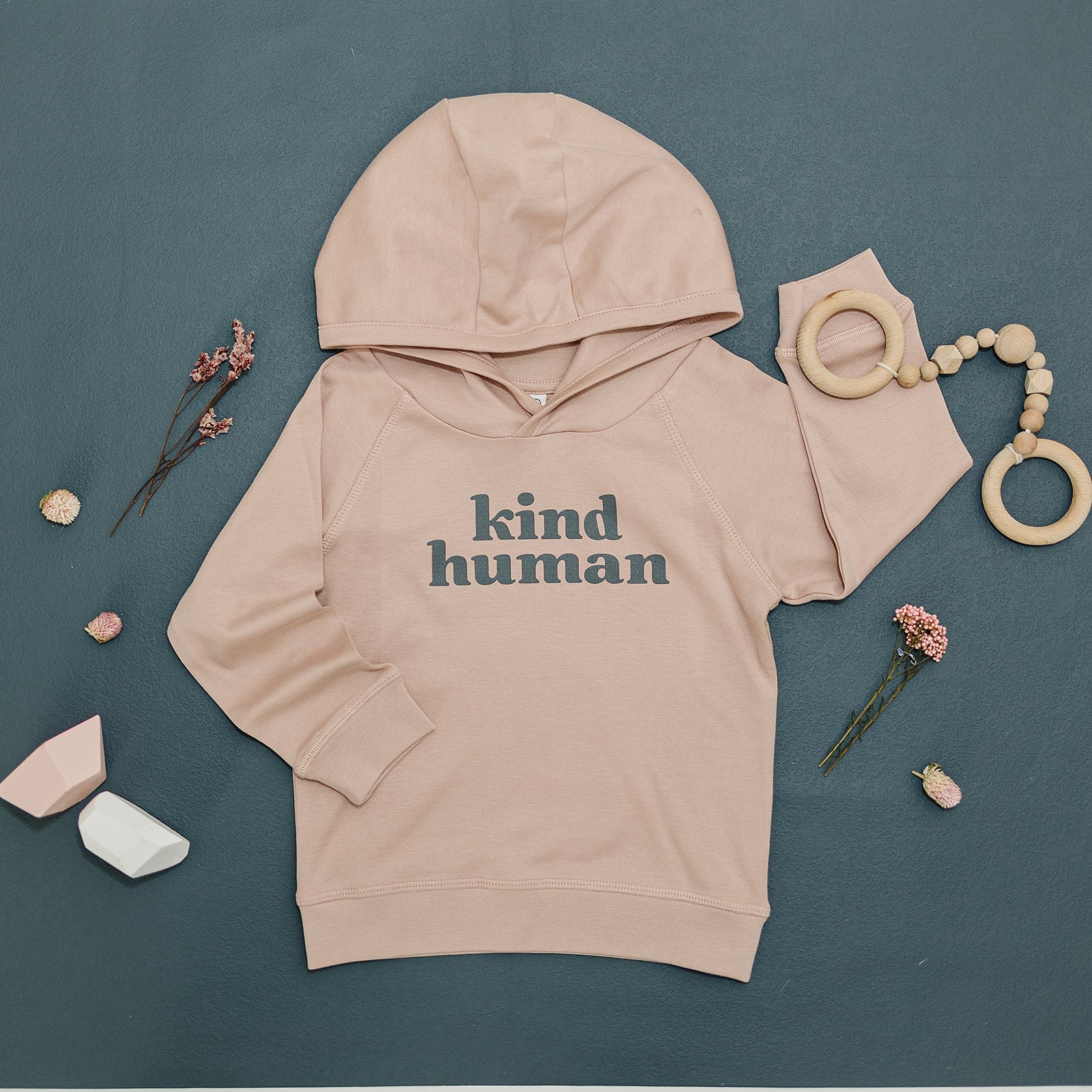Soft beige 'kind human' hoodie laid flat on a dark teal surface, surrounded by whimsical dried flowers and natural wooden baby toys.