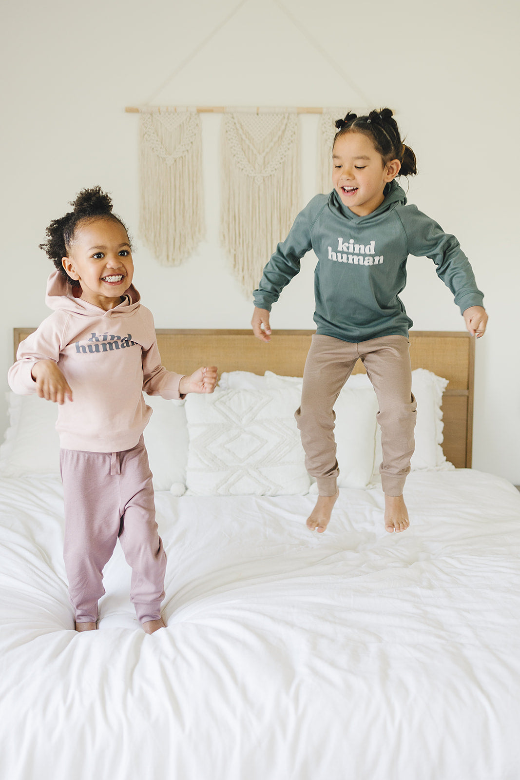 Two joyful children jumping on a bed, one wearing a pink 'kind human' hoodie with matching pants, and the other in an olive hoodie with taupe pants