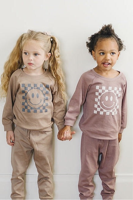 Two smiling children holding hands, one with curly hair and one with straight hair, both wearing taupe sweatshirts with checkered smiley faces and matching pants