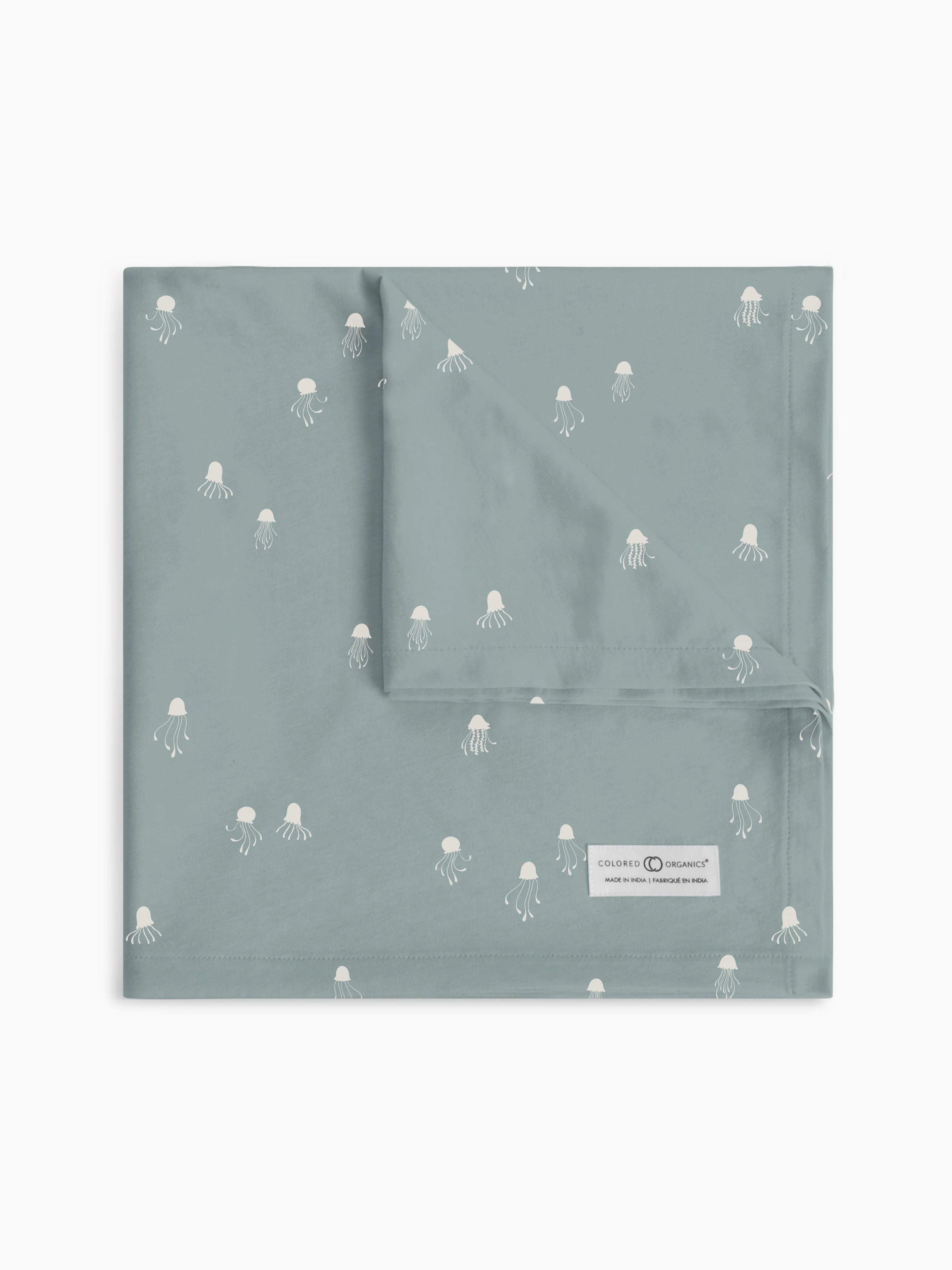 Colored Organics swaddle blanket in 'Jellyfish - Ocean' design, laid flat to show the serene jellyfish pattern on a soft grey background, with the brand's organic commitment label visible