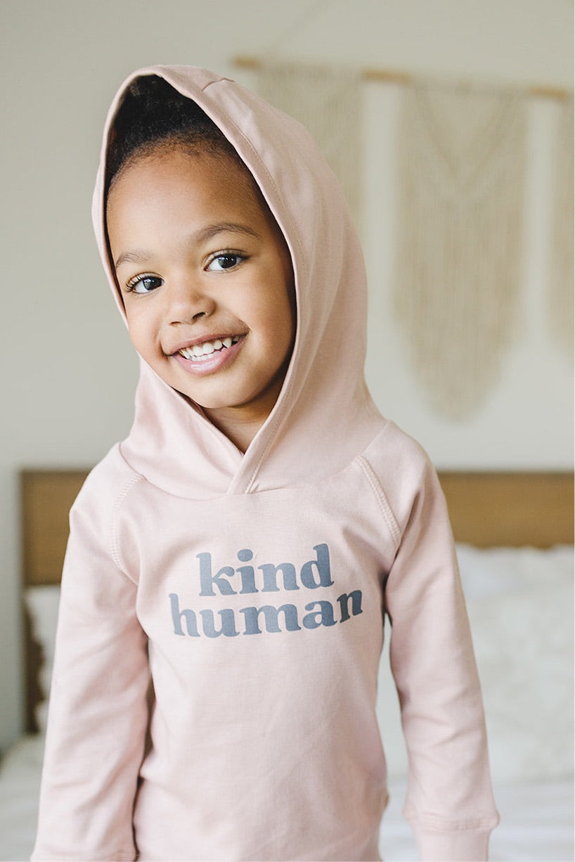 A smiling young child wearing a rose-colored 'Kind Human' hoodie with the hood up
