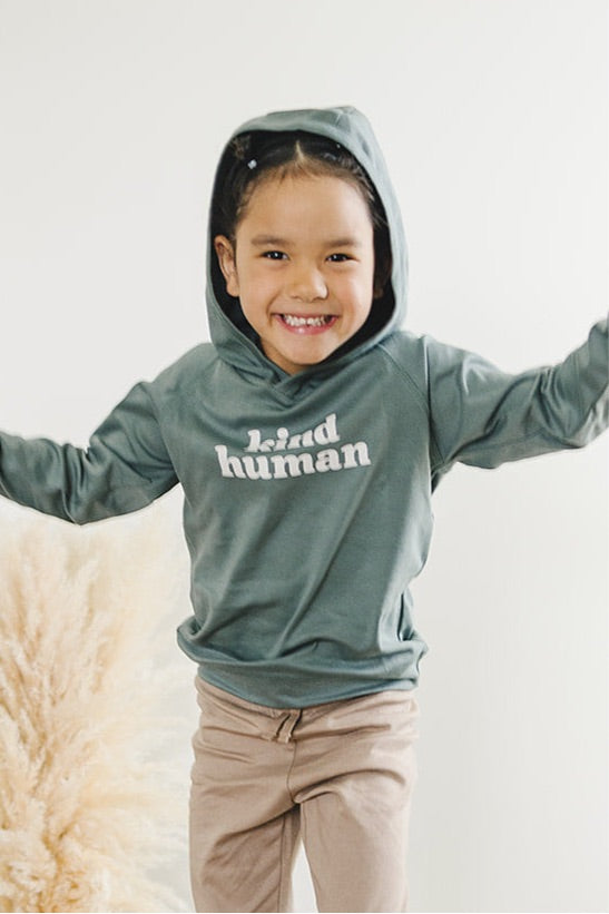 A cheerful child with a beaming smile wearing a sage 'Kind Human' hoodie and beige pants