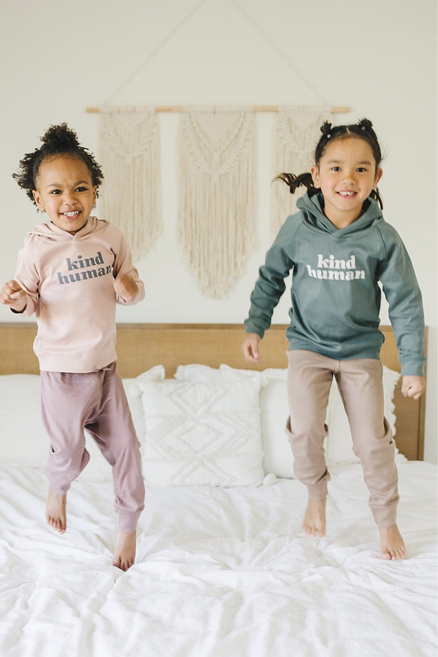 Two joyful children jumping on the bed wearing 'Kind Human' hoodies in pink and sage colors with matching pants.