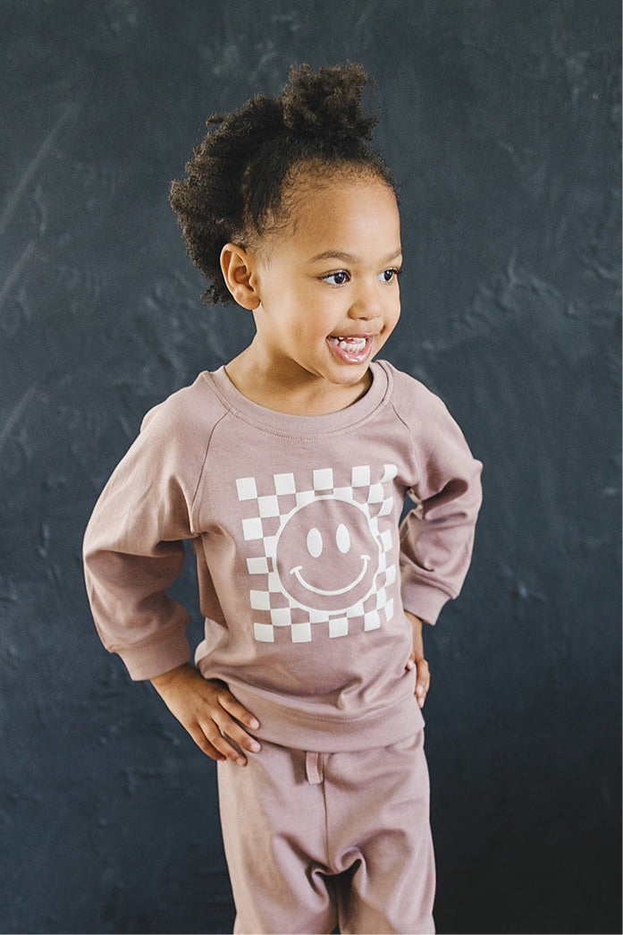 Delighted little girl with her hands on her hips, wearing a taupe sweatshirt with a white checkered smiley face, standing against a textured black background