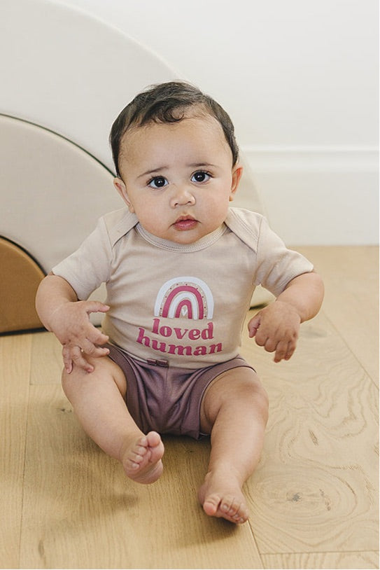 Inquisitive baby sitting on a wooden floor in a neutral-toned 'loved human' onesie with burgundy shorts, with a soft white background