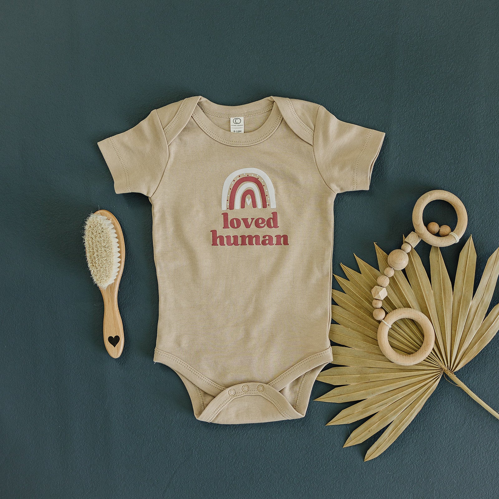Neutral-toned baby onesie with 'loved human' and rainbow print, alongside a natural bristle brush and wooden teething rings on a dark teal background
