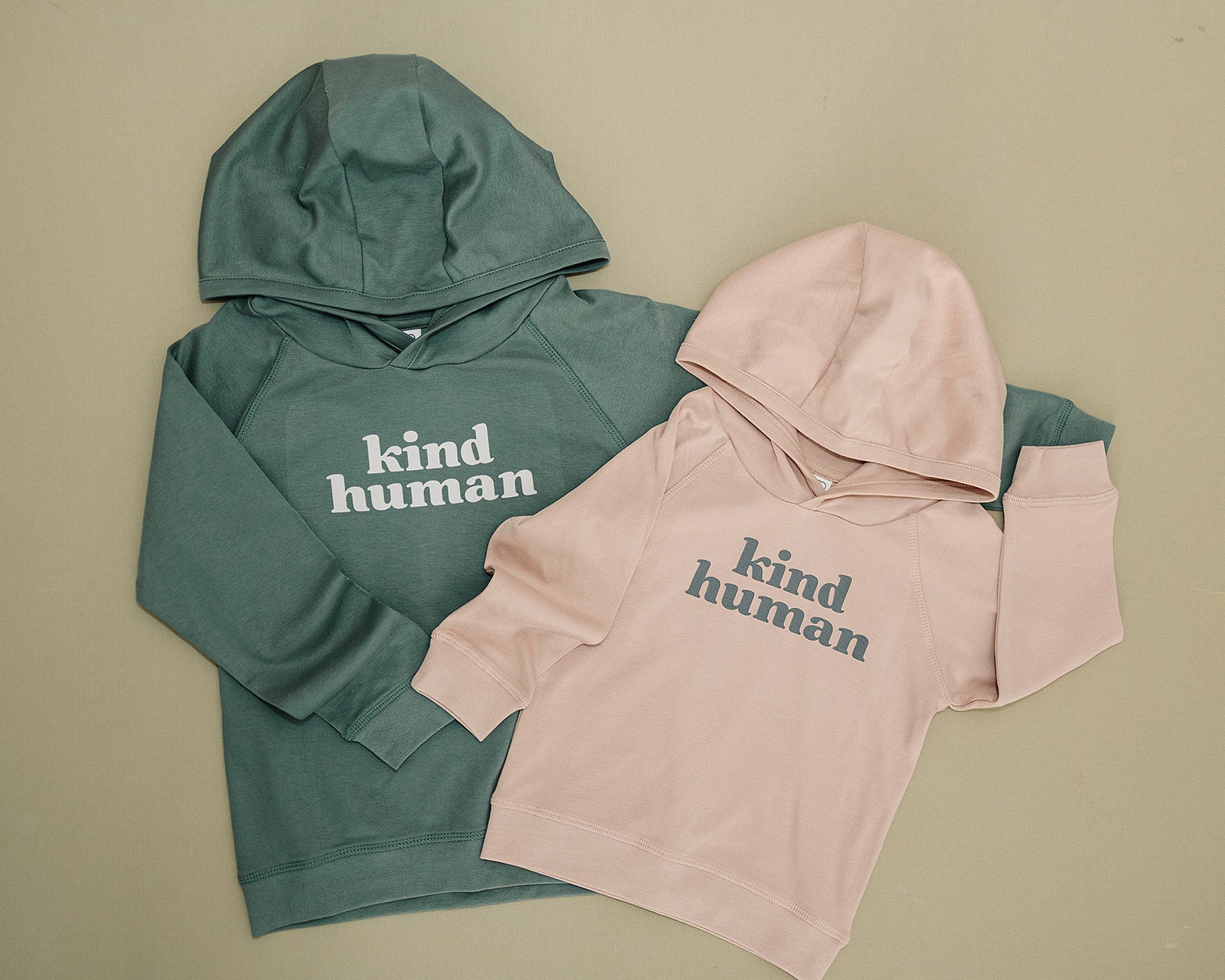 Olive green and blush pink 'kind human' hoodies laid flat on a sage background with a cozy and minimalist vibe