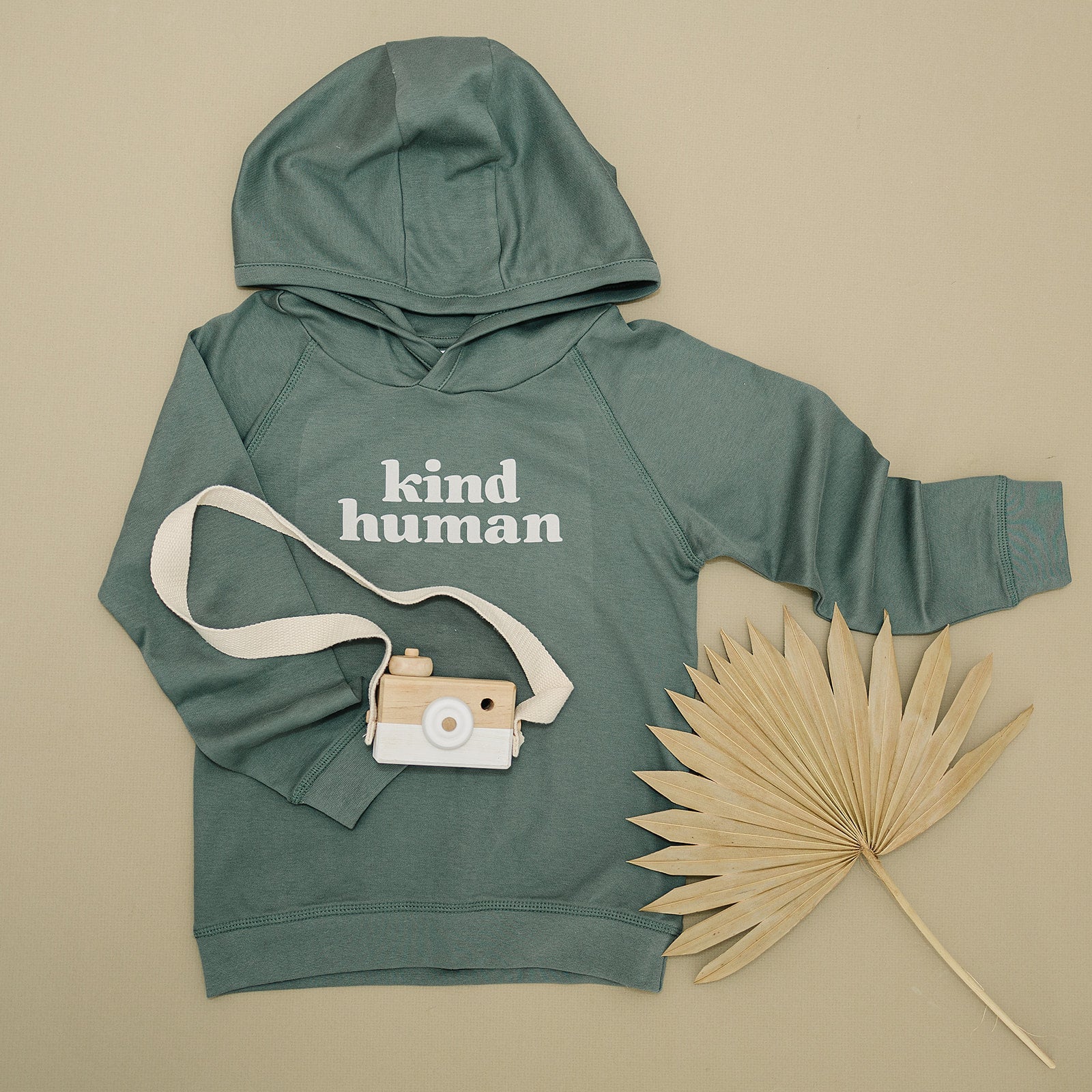 Olive green 'kind human' hoodie laid out with a wooden toy camera and a large dried palm leaf on a beige surface