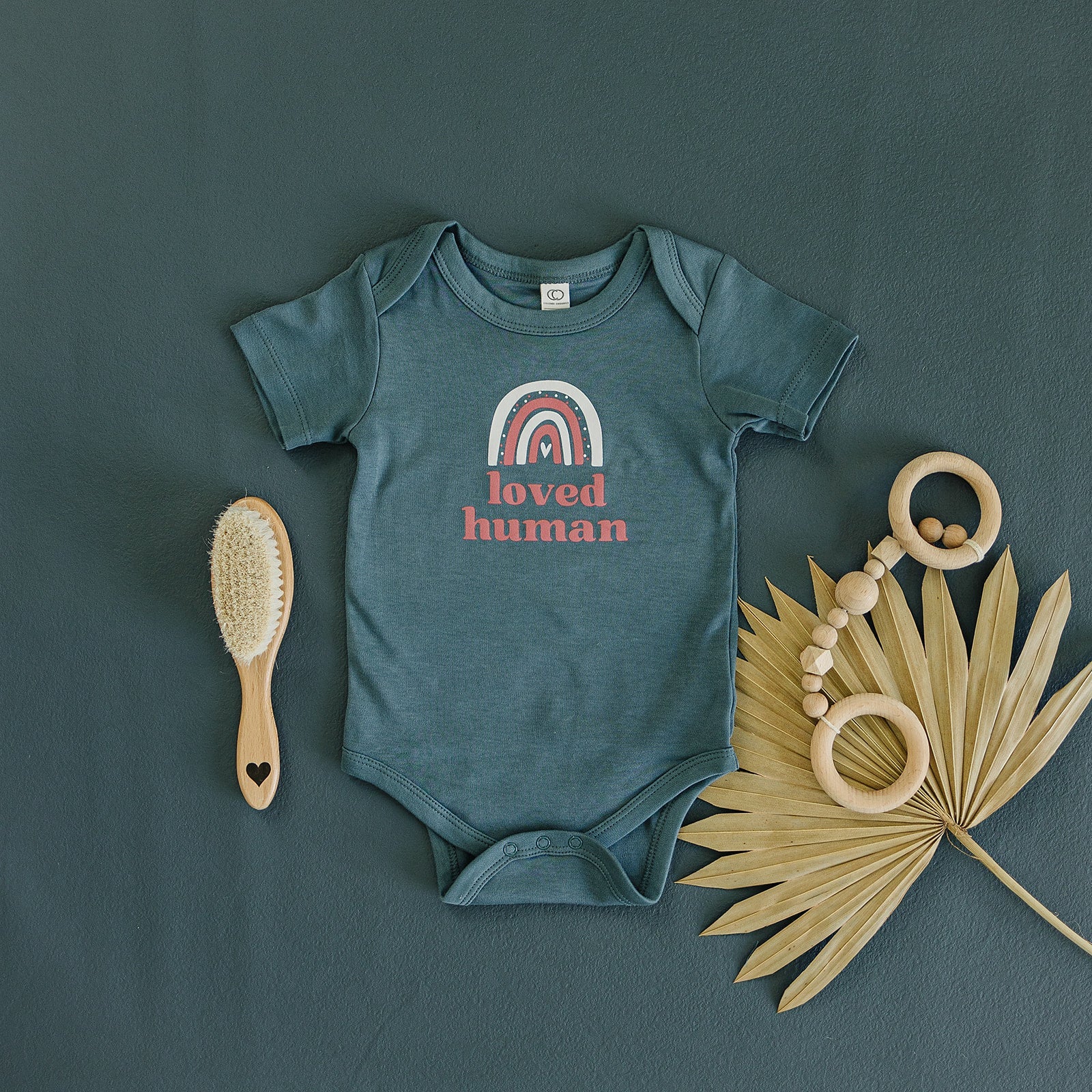 Slate grey baby onesie with 'loved human' and rainbow print, accompanied by a natural bristle brush and wooden teething toys on a dark teal background