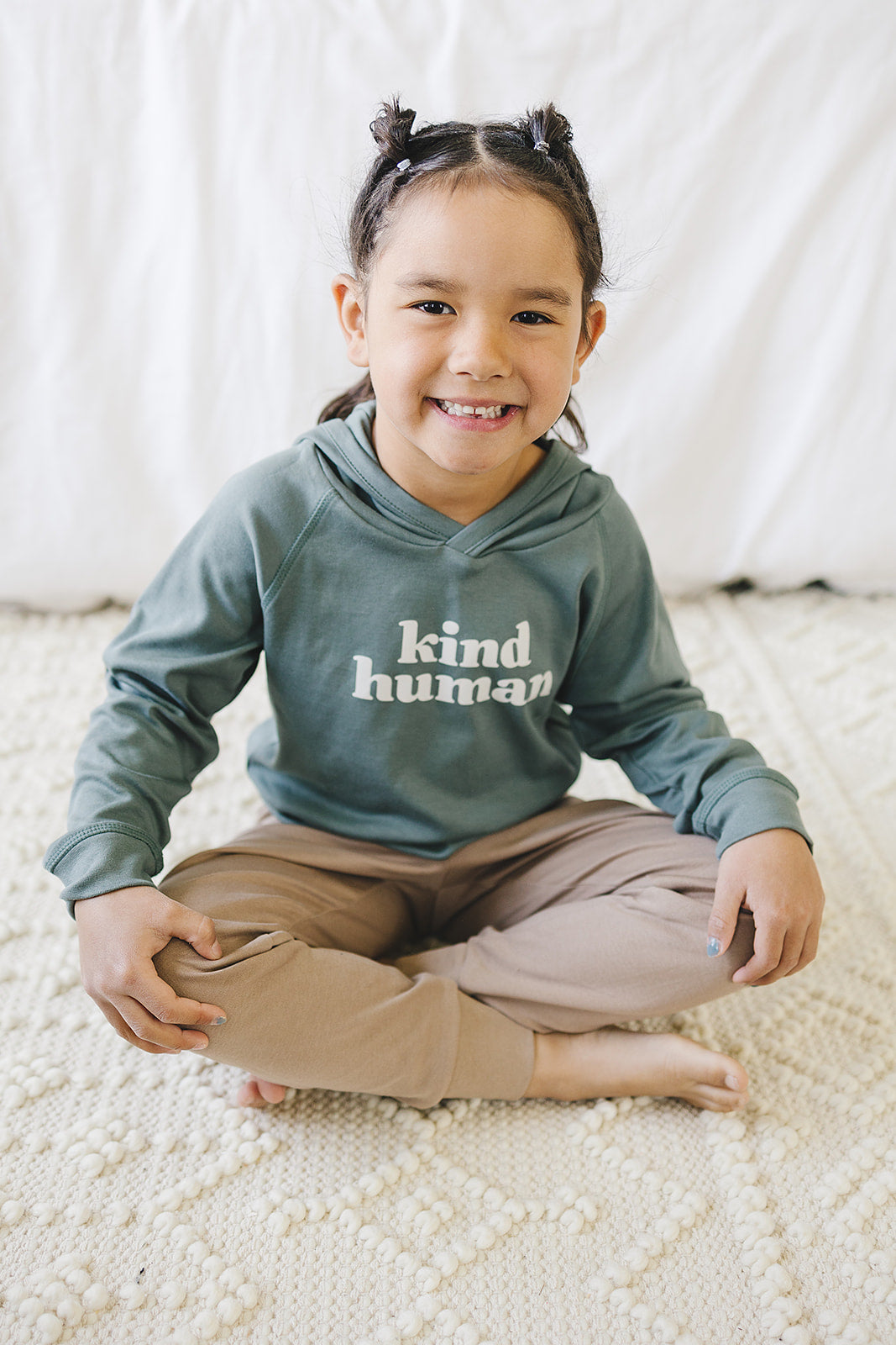 Happy child with a bright smile sitting cross-legged on a textured blanket, wearing an olive green 'kind human' hoodie and taupe pants