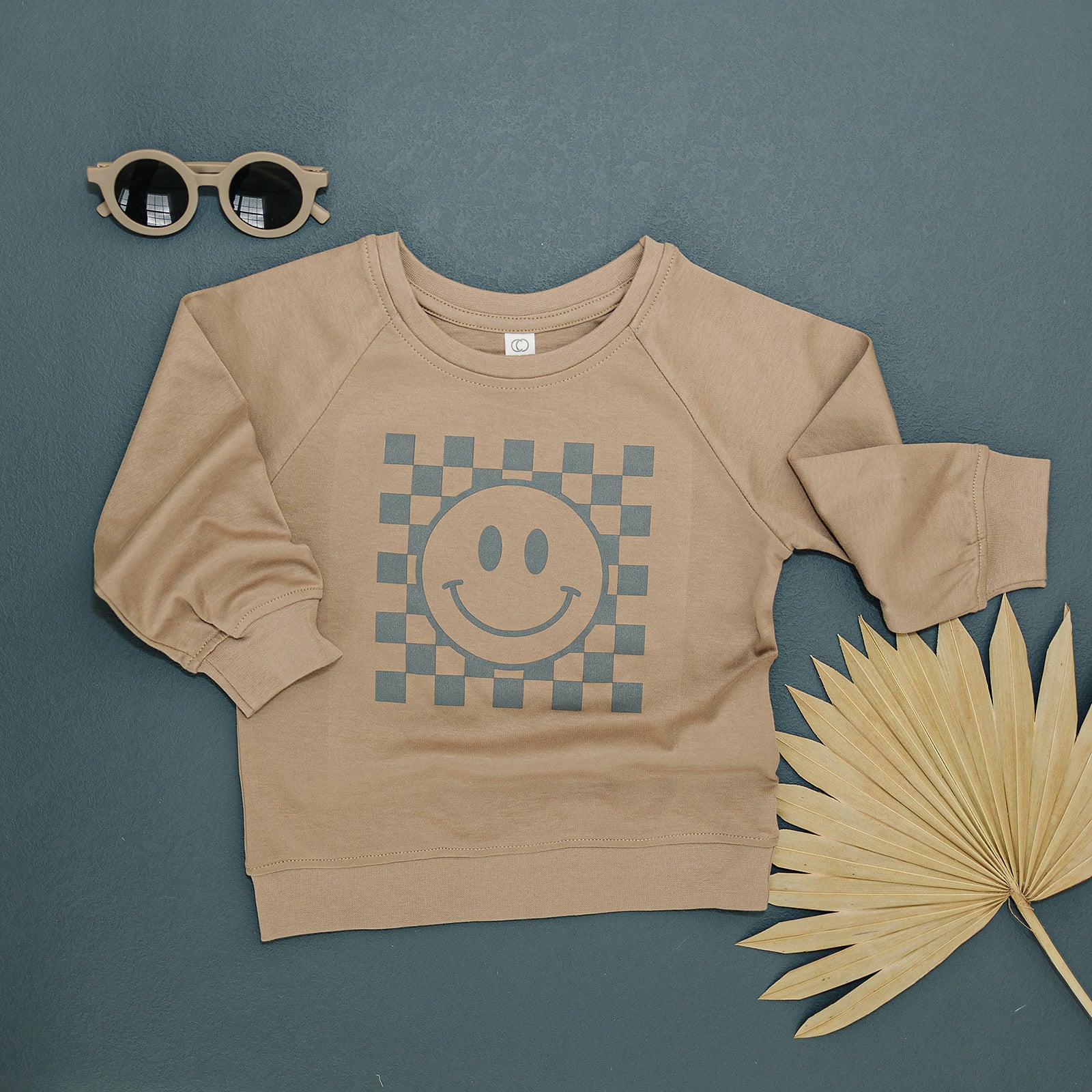 Stylish tan crewneck sweatshirt with a checkered smiley face design, displayed with a pair of round sunglasses and a dried palm fan on a dark grey background