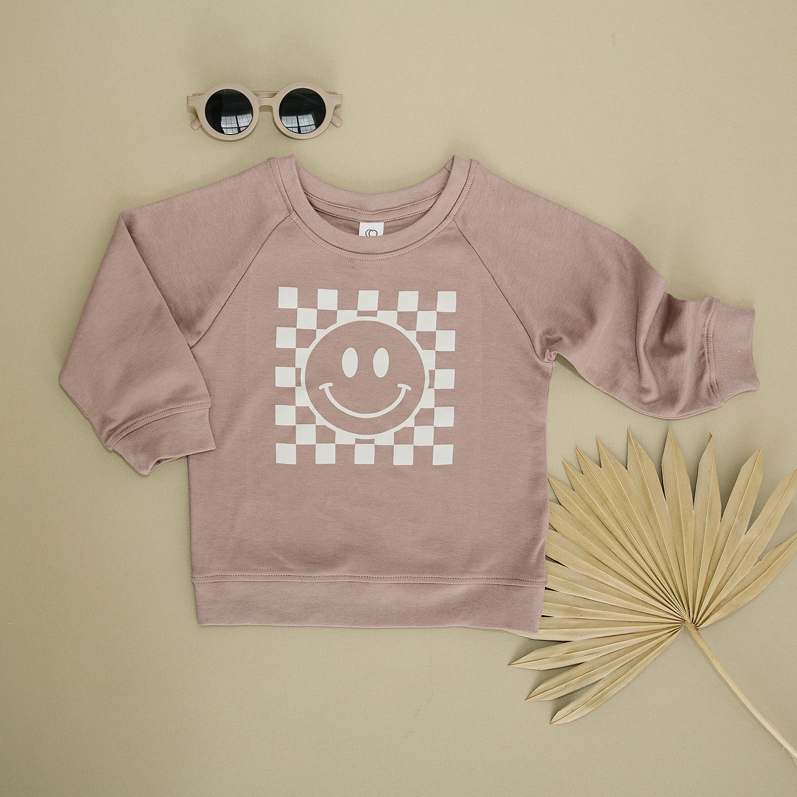 Chic taupe children’s sweatshirt featuring a white checkered smiley face, displayed with a pair of round sunglasses and a dried palm leaf on a khaki background