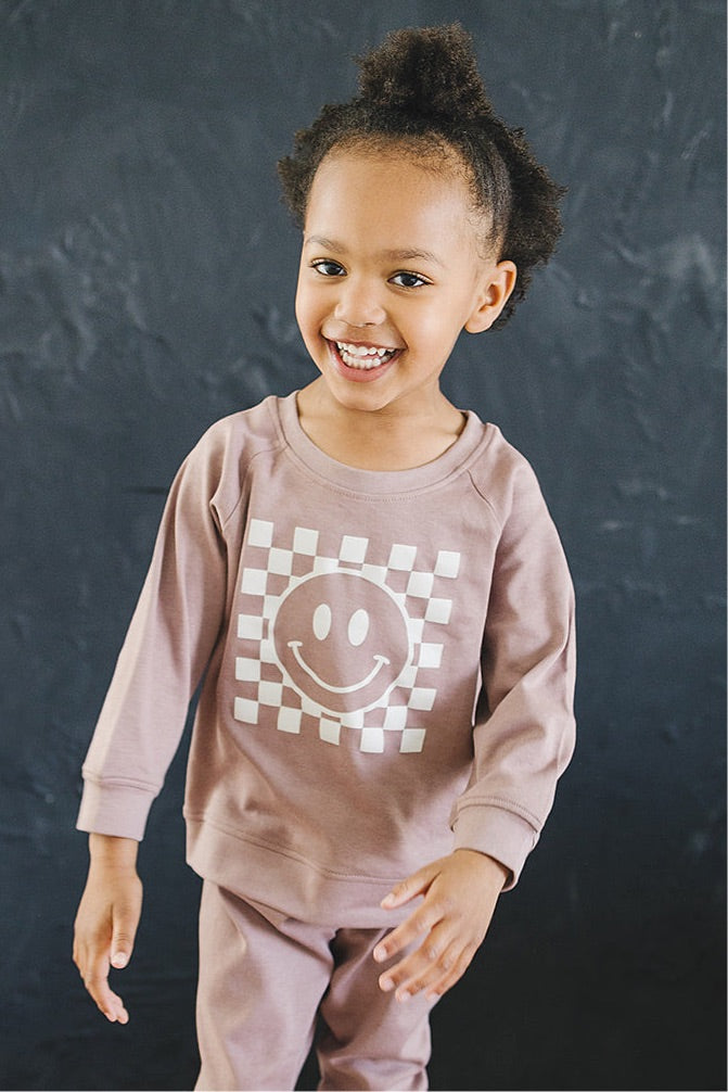 Cheerful young girl posing in a cozy taupe sweatshirt with a checkered smiley face design, paired with matching pants against a dark background